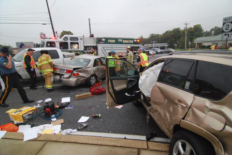 Emergency responders at the scene of a multi-car collision on a busy road. Several vehicles are damaged, debris is scattered, and a fire rescue truck is present. Traffic is stopped, and responders are assisting and assessing the situation.