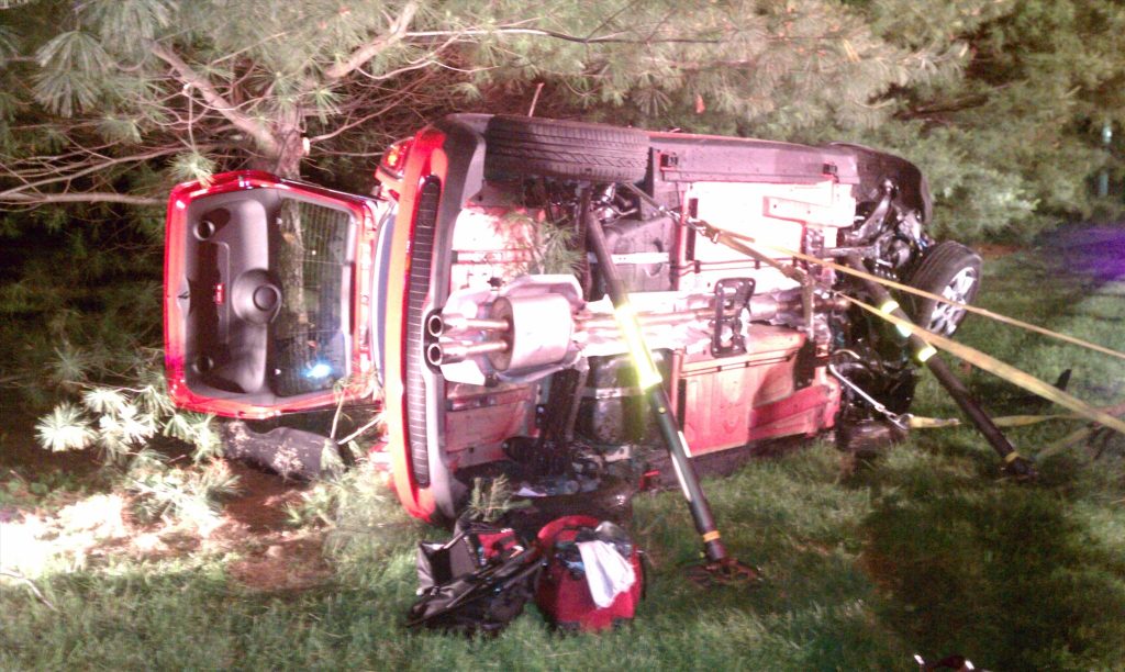 A red SUV is overturned on its side on a grassy area near pine trees. The undercarriage and exhaust system are visible. Various emergency tools and equipment are scattered around the scene.