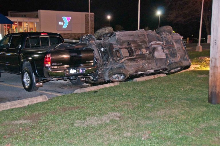 A black pickup truck and another vehicle are involved in an accident in a parking lot at night. The second vehicle is overturned, leaning on the pickup. A lit building with a YMCA logo is visible in the background.