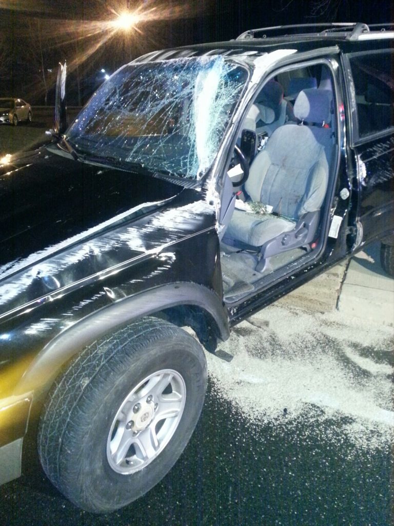 A damaged black SUV with a shattered windshield and the driver’s side door open. Debris is scattered on the ground next to the vehicle. It is nighttime, and a streetlight is visible in the background.