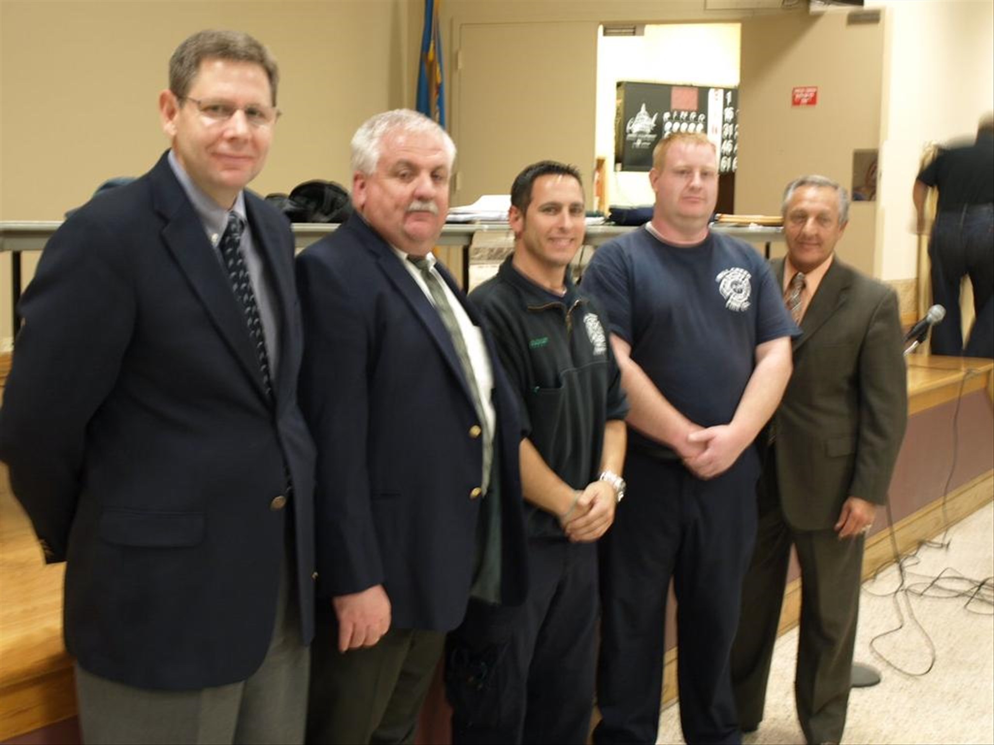 Five men stand indoors, wearing suits and uniforms. They are positioned in a line facing the camera, appearing formal.