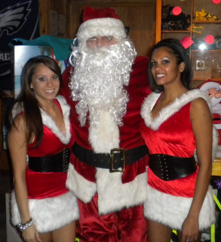 Santa Claus stands smiling between two women dressed in matching red and white Christmas outfits. They are inside a room with festive decorations, creating a cheerful holiday atmosphere.