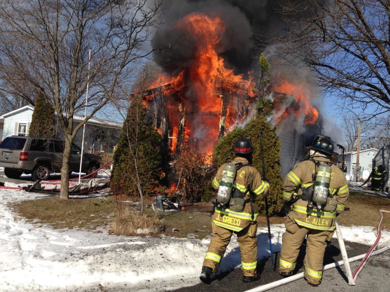 Two firefighters in yellow gear and helmets face a burning house engulfed in flames and thick smoke. Snow is on the ground, and bare trees surround the scene. A parked SUV is visible on the left.