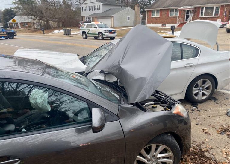 Two cars involved in a front-end collision on a suburban street. A dark gray car has collided with a white car. Both vehicles have popped hoods, and debris is visible on the road. Emergency vehicle in the background.