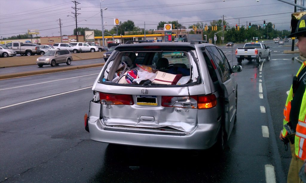 A silver minivan with a crumpled rear end is stopped on a wet road. The back is filled with belongings. A firefighter in uniform stands nearby. Cars are in the background near a gas station and traffic lights. It's a cloudy day.