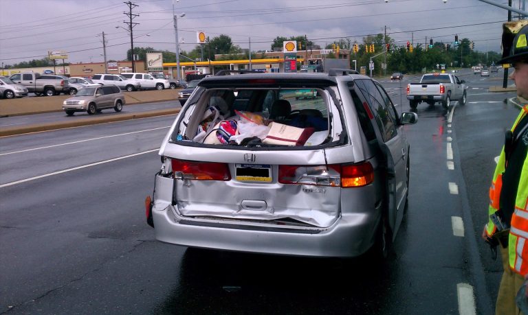 A silver minivan with a crumpled rear end is stopped on a wet road. The back is filled with belongings. A firefighter in uniform stands nearby. Cars are in the background near a gas station and traffic lights. It's a cloudy day.