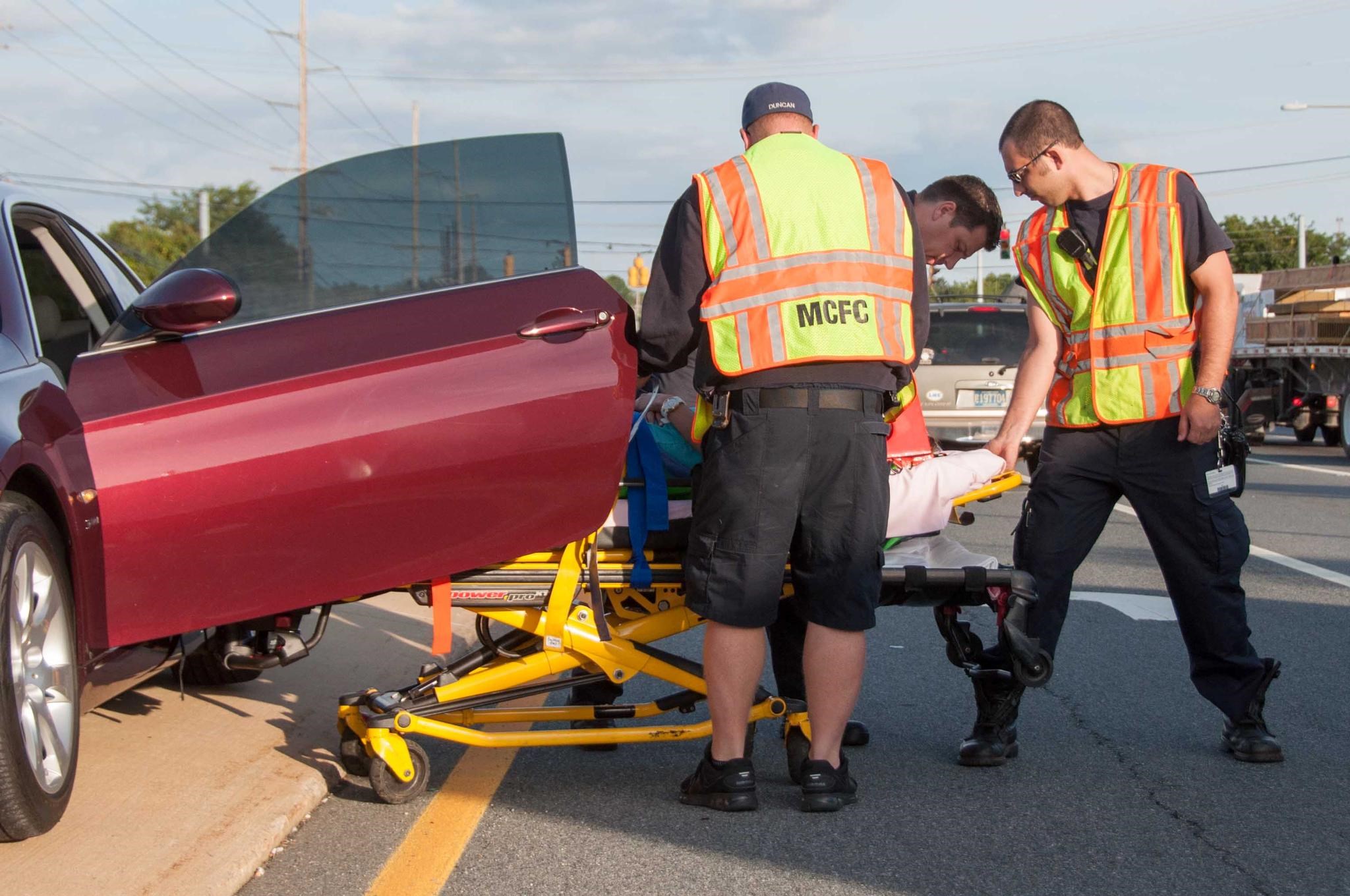 Three emergency responders in high-visibility vests assist a person on a stretcher near a maroon car parked beside a road. The responders are focused and appear to be preparing to move the individual.