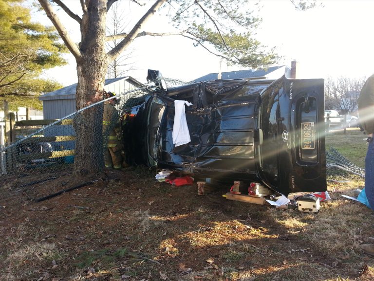 A black car is flipped on its side against a tree and a chain-link fence. A firefighter in yellow gear is inspecting the scene. Debris and tools are scattered on the grass around the vehicle. Trees and a building are in the background.