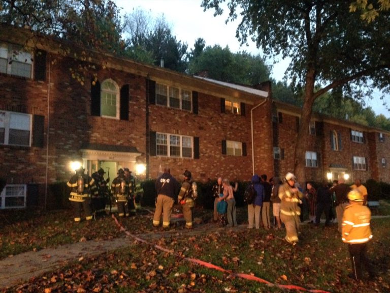 Firefighters in uniform gather outside a brick apartment building at dusk, with autumn leaves on the ground. A group of people stands nearby, some holding bags. Red tape is visible in the foreground.