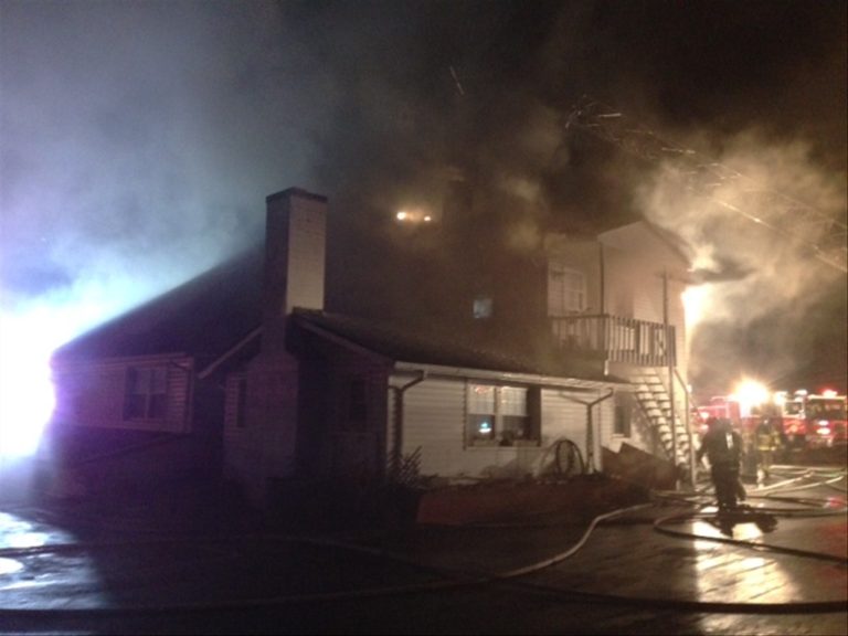 A nighttime scene of a two-story house on fire, with smoke billowing from the roof. Firefighters are present, spraying water hoses towards the building. Emergency vehicle lights illuminate the area.