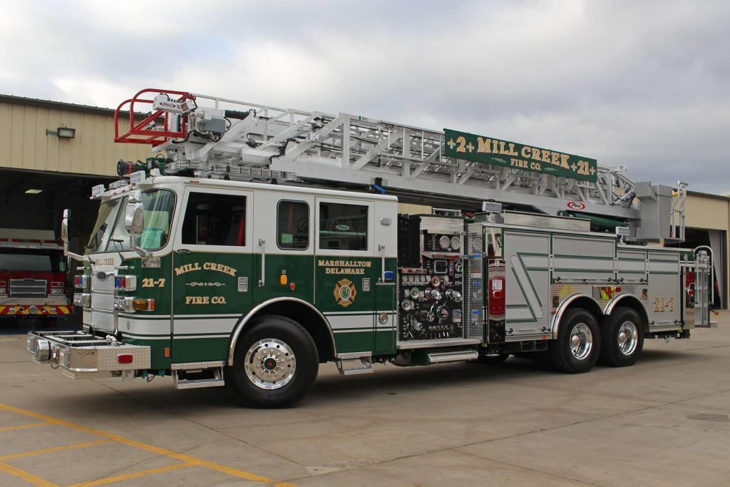 A fire truck labeled "Mill Creek" is parked outside a garage. It has an extendable ladder on top and multiple storage compartments on the sides. The truck is white with green and gold accents.