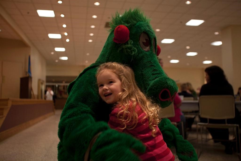 A young girl with curly blonde hair, wearing a pink striped shirt, happily hugs a person in a green dragon costume inside a large room with people sitting at tables in the background.