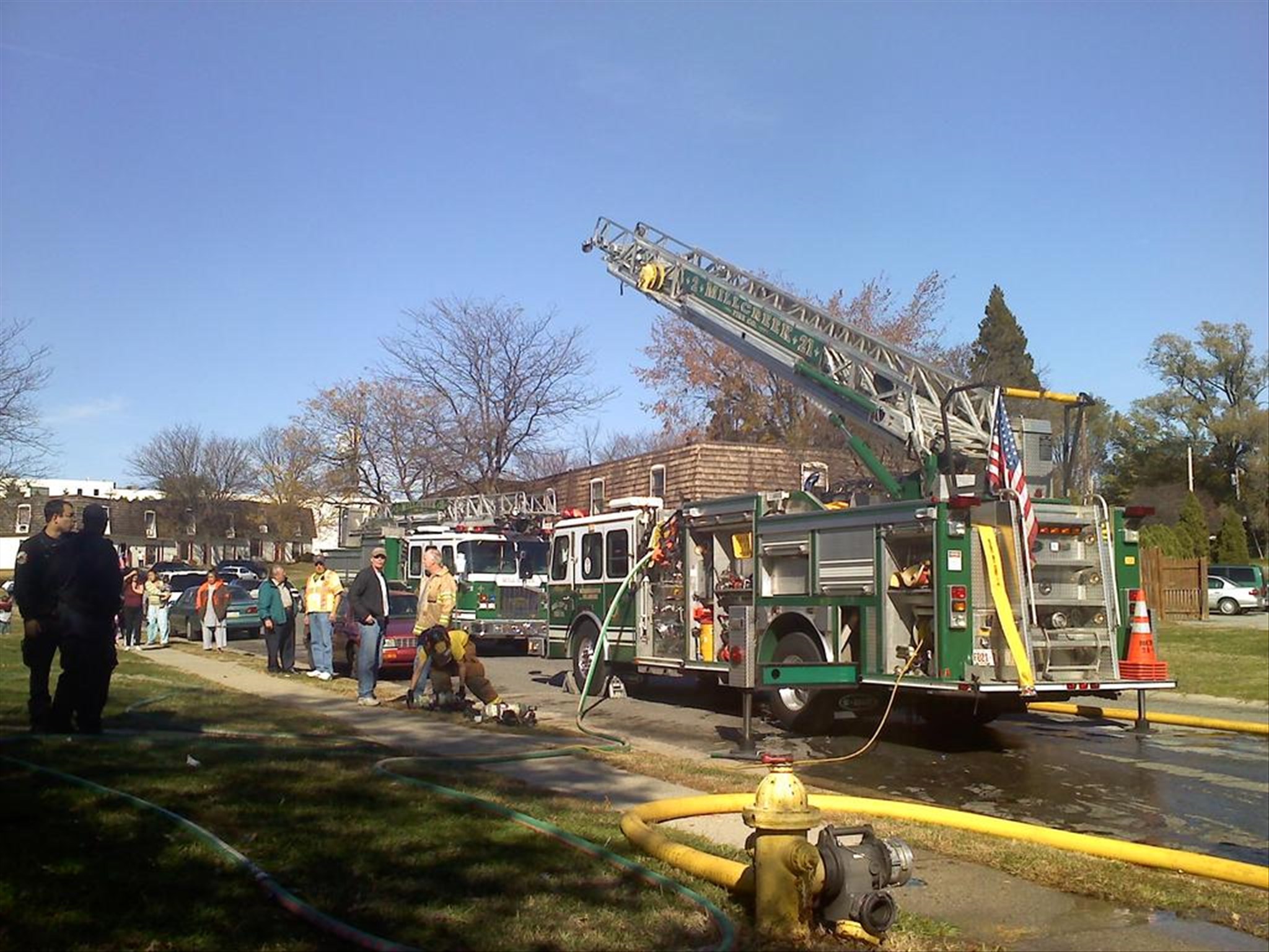 A fire truck with a raised ladder is parked on a street. Firefighters and bystanders are nearby, with hoses extending from the truck. Buildings and trees are visible in the background under a clear blue sky.