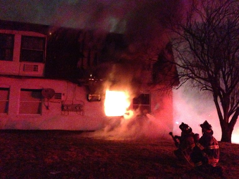 Firefighters spray water at a blazing fire in a two-story building at night. Flames and thick smoke pour from a window on the upper floor. Nearby, a leafless tree stands silhouetted against the illuminated smoke.
