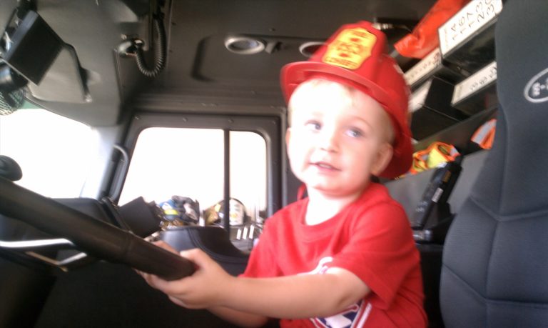 A young child wearing a red fireman's helmet is sitting in the driver's seat of a fire truck, holding the steering wheel. He is smiling and dressed in a red shirt. The truck interior has various equipment visible.