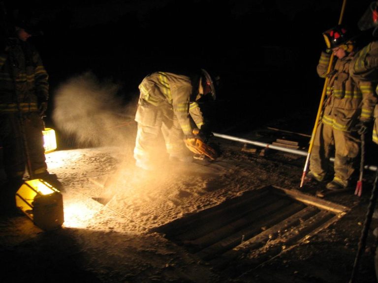 Firefighters work at night using a circular saw to cut through a roof, with dust illuminated by the light. Other firefighters stand nearby, holding equipment and overseeing the operation.