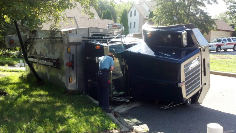 A large armored truck is overturned on its side on a grassy area by a road. A police officer is inspecting the vehicle. Emergency vehicles are visible in the background. Trees and residential buildings are nearby.