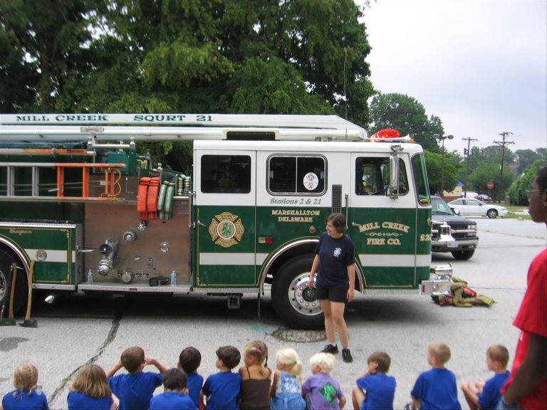 A group of children sits on the ground, watching a firefighter standing in front of a green and white fire truck labeled "MILL CREEK SQUAD 21." The truck is parked outdoors near trees, and some adults are standing nearby.