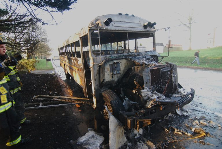 A burned-out school bus with severe fire damage is parked on the side of the road. The front is completely charred, and debris is scattered around. A firefighter is on the left, and a person walks in the background on the right.