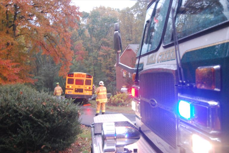 A fire truck with flashing lights is parked on a suburban street. Firefighters in yellow suits stand nearby. A yellow school bus is visible in the background, surrounded by autumn trees and a residential house.