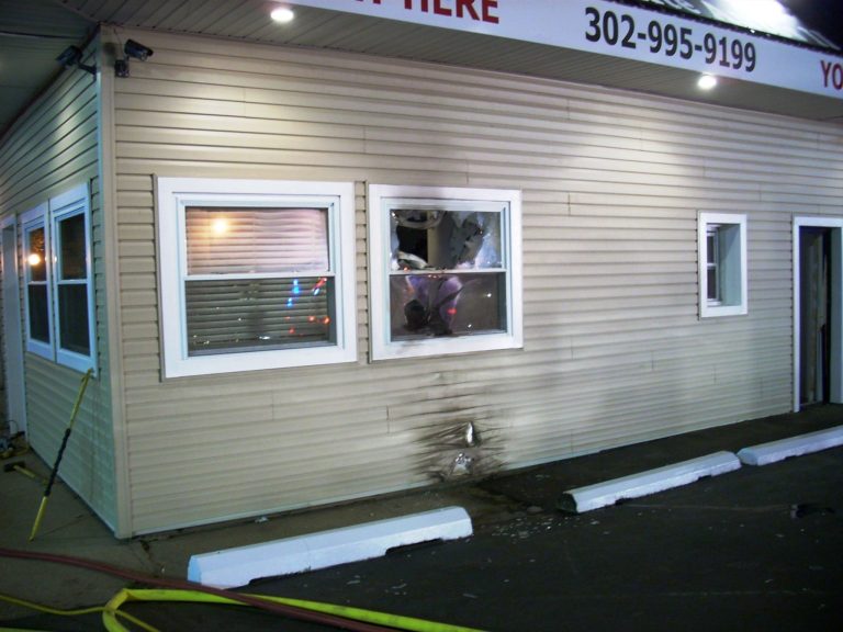 A beige building with visible fire damage below a broken window. The siding is scorched, and there's debris on the window ledge. Yellow hoses are on the ground, and a parking curb is nearby. Overhead lights illuminate the scene.