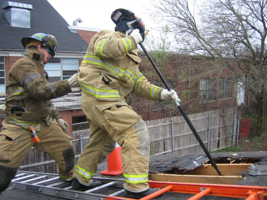 Two firefighters in full gear are on a roof. One uses a tool to open a hole, while the other looks on. A ladder is visible, and a tree with sparse leaves is in the background.
