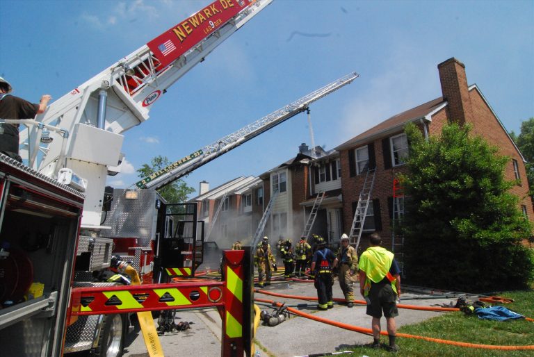 Firefighters respond to a house fire at a brick townhouse. A fire truck with an extended ladder is visible, and several firefighters in gear are actively working with hoses. Smoke is coming from the building, and the scene is busy with emergency personnel.