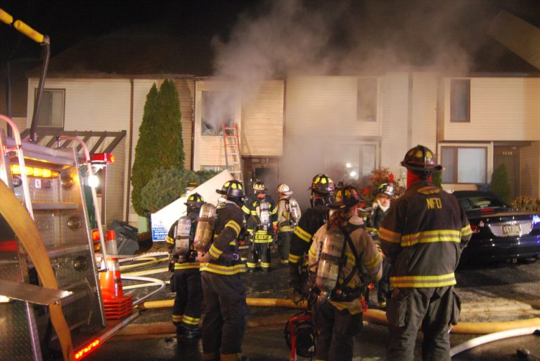 Firefighters stand outside a smoking residential building at night. They are wearing protective gear and helmets. Fire hoses are on the ground, and a fire truck is visible on the left. Smoke is coming from a window in the building.