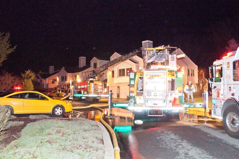 Fire trucks and emergency personnel respond to a nighttime scene outside a large residential building. A yellow car is parked on the street, and hoses are laid out, indicating firefighting activity. The area is well-lit by the trucks' lights.