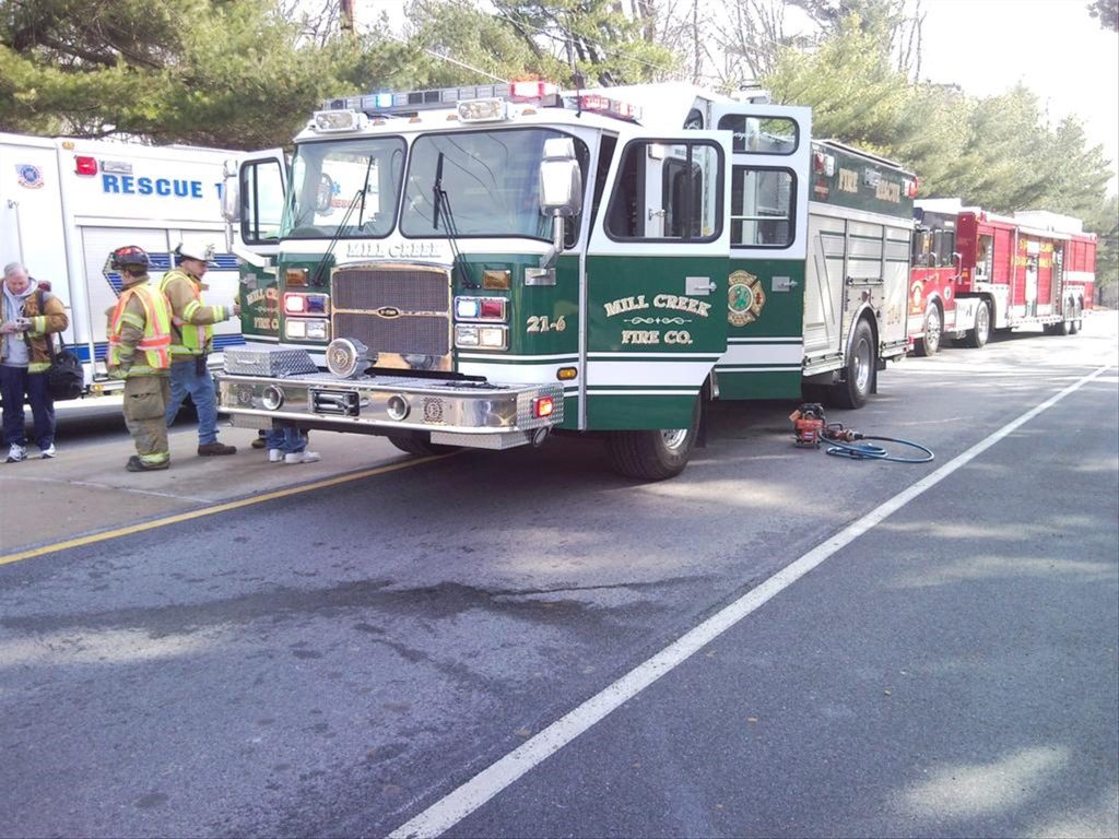 A green and white fire truck with "Mill Creek Fire Co." on the side is parked on the road. Firefighters in uniform stand nearby. Other emergency vehicles and trees line the background. Equipment is placed on the ground next to the truck.
