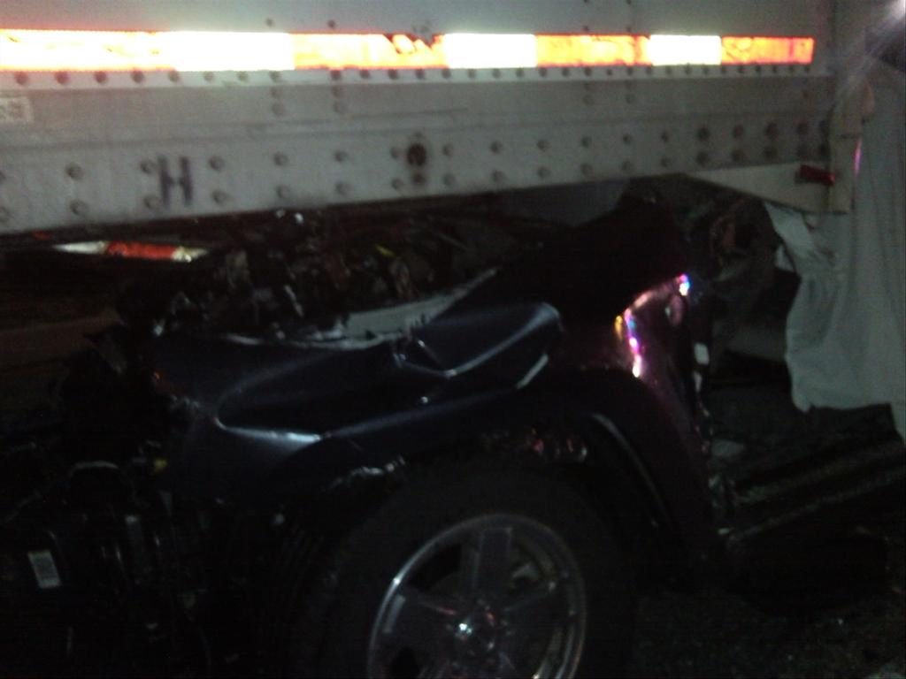 A car is partially lodged under a large truck trailer. The car's front is heavily damaged and crumpled. The image is dimly lit, highlighting the extent of the collision with reflective sections on the trailer.