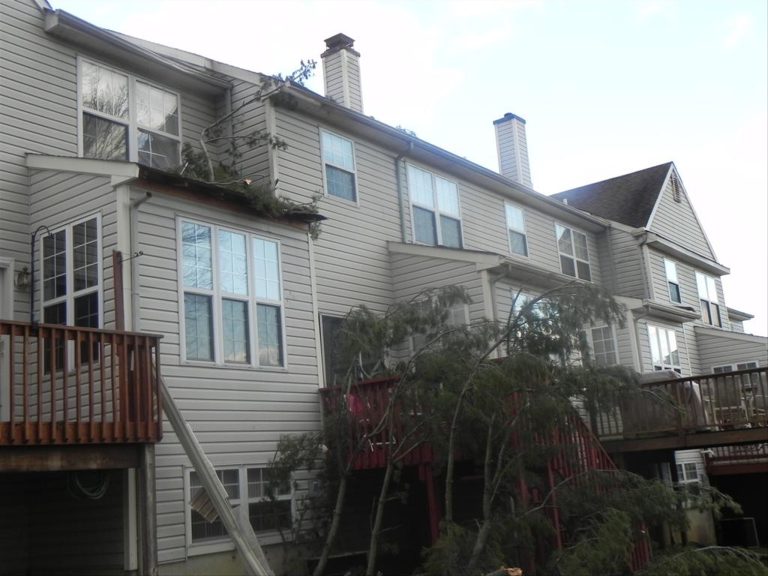 A large tree has fallen onto the roof and second-story balcony of a row of attached townhouse buildings, causing visible damage. The buildings have beige siding, with multiple windows and chimneys. The sky is lightly overcast.