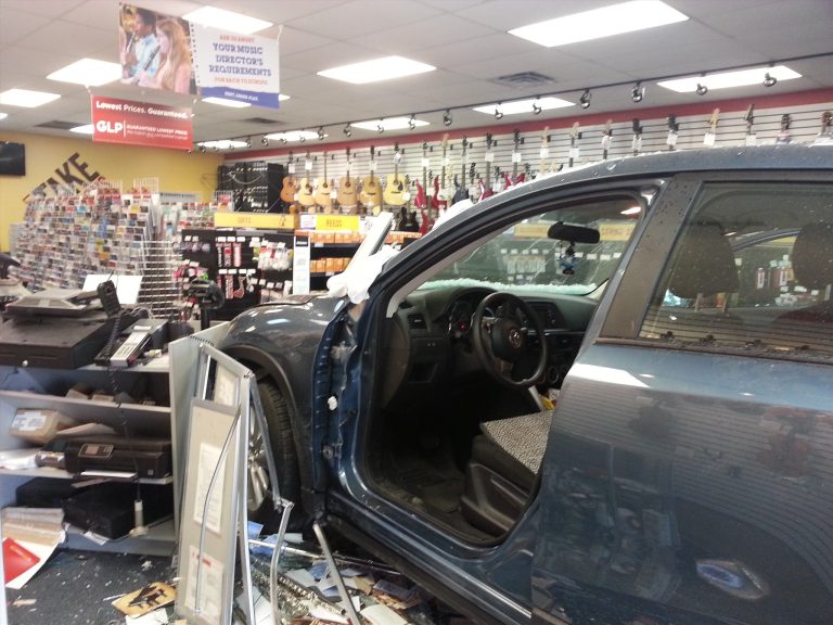A car has crashed into a music store, stopping inside near shelves of guitars and music equipment. The driver's side door is open, and debris is scattered on the floor. The store's interior and various instruments are visible.