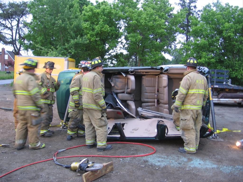 Firefighters in gear assess a vehicle on its side during a rescue operation. The scene includes trees in the background, a yellow barricade, and tools on the ground.