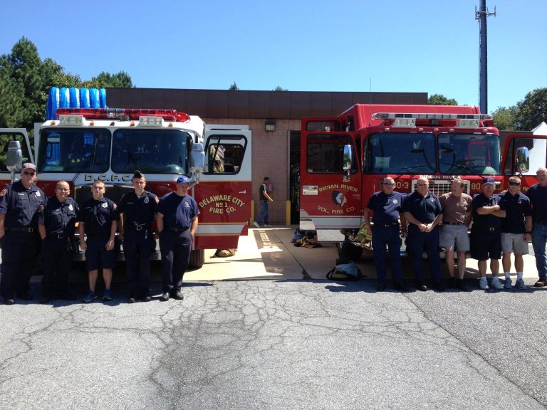 A group of firefighters stands next to two fire trucks in front of a fire station. One fire truck is labeled "Delaware City Fire Co." and the other "Indian River Vol. Fire Co." The firefighters are wearing uniforms and looking towards the camera.