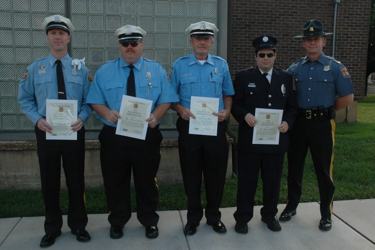 Five uniformed police officers stand in a row outdoors, four holding certificates. They are in front of a brick building with glass block windows. The officers wear dark pants; three have light blue shirts, one has a black shirt, and one has a dark blue uniform.