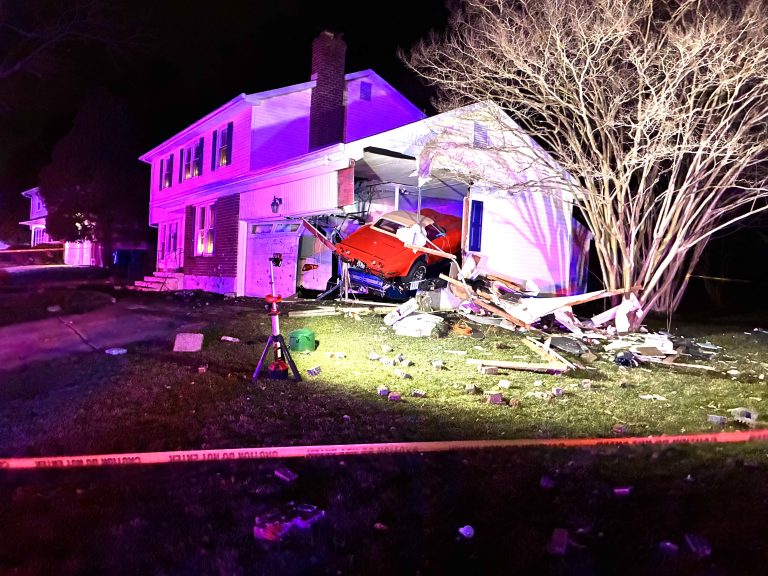 A red car has crashed into the side of a house, partially embedded in the structure. Debris is scattered around the yard. The scene is lit with purple and white lights. Yellow caution tape is visible in the foreground.
