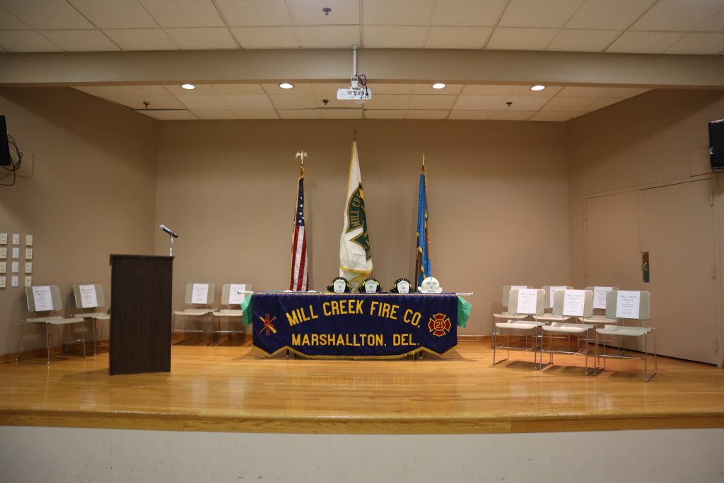 A stage with a podium, U.S. and state flags, and a table covered with a "Mill Creek Fire Co., Marshallton, Del." banner, displaying helmets and awards. Empty chairs with name placards line both sides.