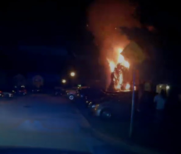 A large fire burns intensely near parked cars at night, sending flames and smoke into the air. Several people stand nearby, watching the blaze. The scene is illuminated by the fire and nearby streetlights.