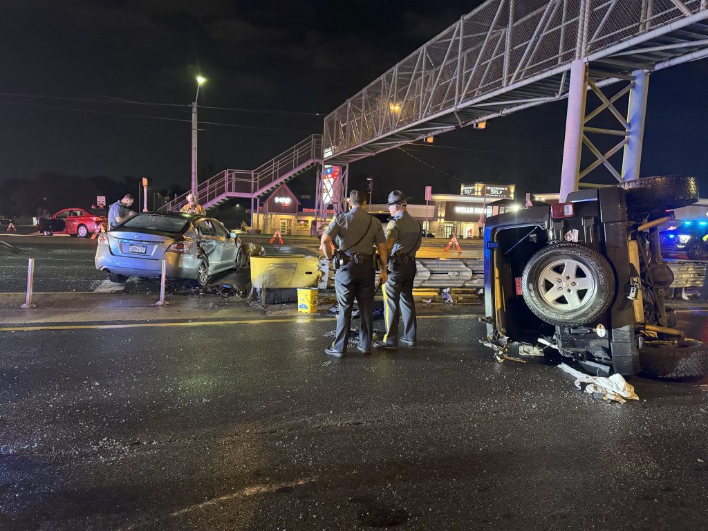 Two police officers stand near an overturned SUV and a damaged car after a nighttime accident beneath a pedestrian bridge, with emergency lights and cones visible.