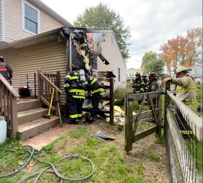 Firefighters in full gear extinguish a fire on the exterior of a beige two-story house, with charred siding and smoke damage visible. Debris is scattered on the ground, and hoses run across the yard.