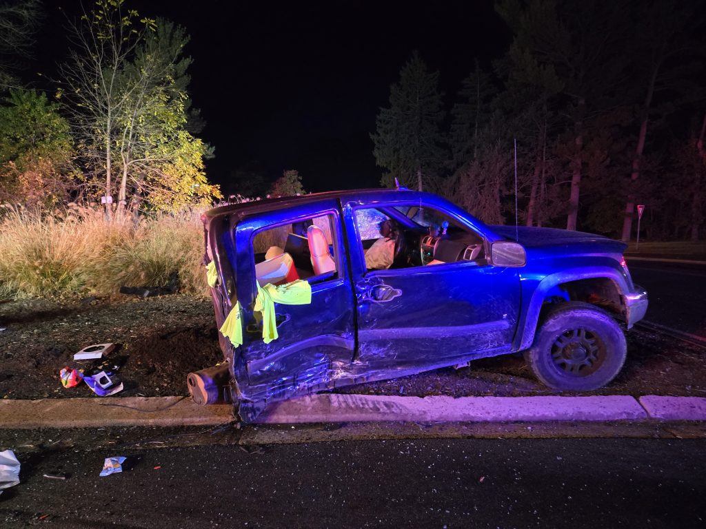 A damaged blue pickup truck with its side smashed rests partially on a curb at night. The driver’s door is missing, and debris is scattered nearby. Trees and grass are visible in the background.