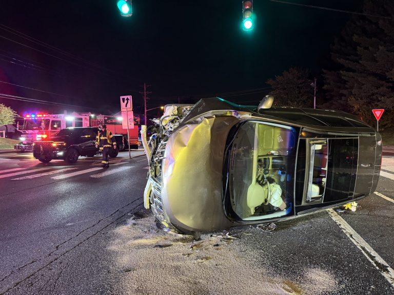 A black SUV lies on its side in the middle of an intersection at night, with visible damage. Emergency responders and vehicles with flashing lights are present nearby under green traffic lights.