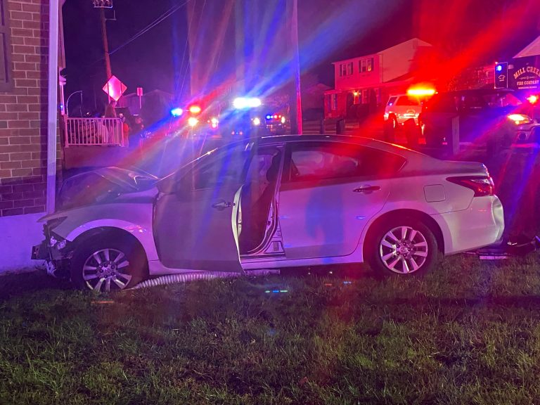 A silver car with its front end crashed into a brick building, driver’s door open. Police vehicles with flashing lights surround the scene at night. Grass and debris are visible in the foreground.