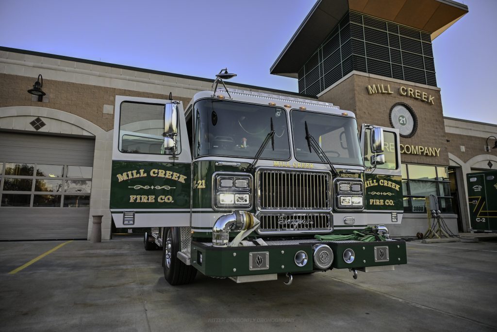 A green and white fire truck labeled "Mill Creek Fire Co." is parked in front of the Mill Creek Fire Company station building on a sunny day.