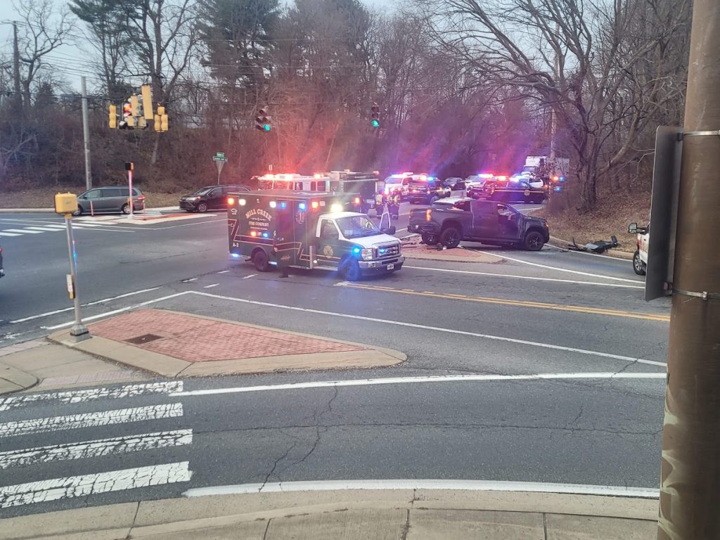 Multiple emergency vehicles with flashing lights block an intersection, including an ambulance, police cars, and a tow truck, with a damaged black car on the side and trees in the background.