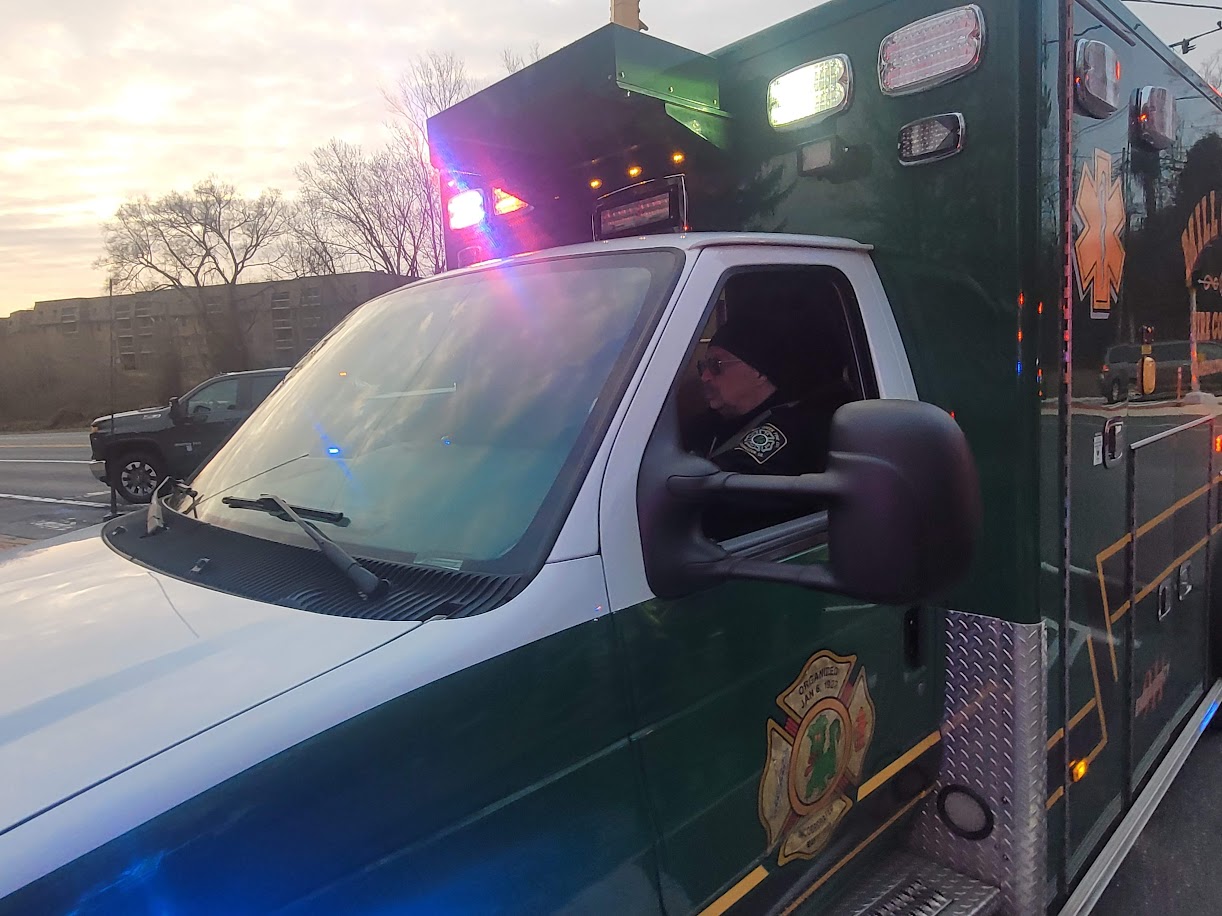 A green and white ambulance with flashing lights is parked on a street. A uniformed person is seated in the driver's seat, and a building and trees are visible in the background during sunset.
