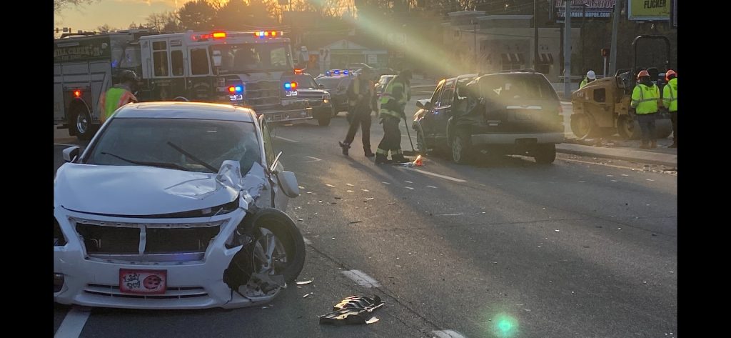 Two damaged vehicles in the foreground after a car accident on a road. Emergency responders, including fire trucks with flashing lights, are on the scene. Workers in reflective vests stand nearby as the sun sets in the background.
