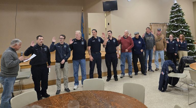 A group of people stands in a line with several raising their right hands, possibly taking an oath, as a man reads from a paper. A Christmas tree is decorated in the background.