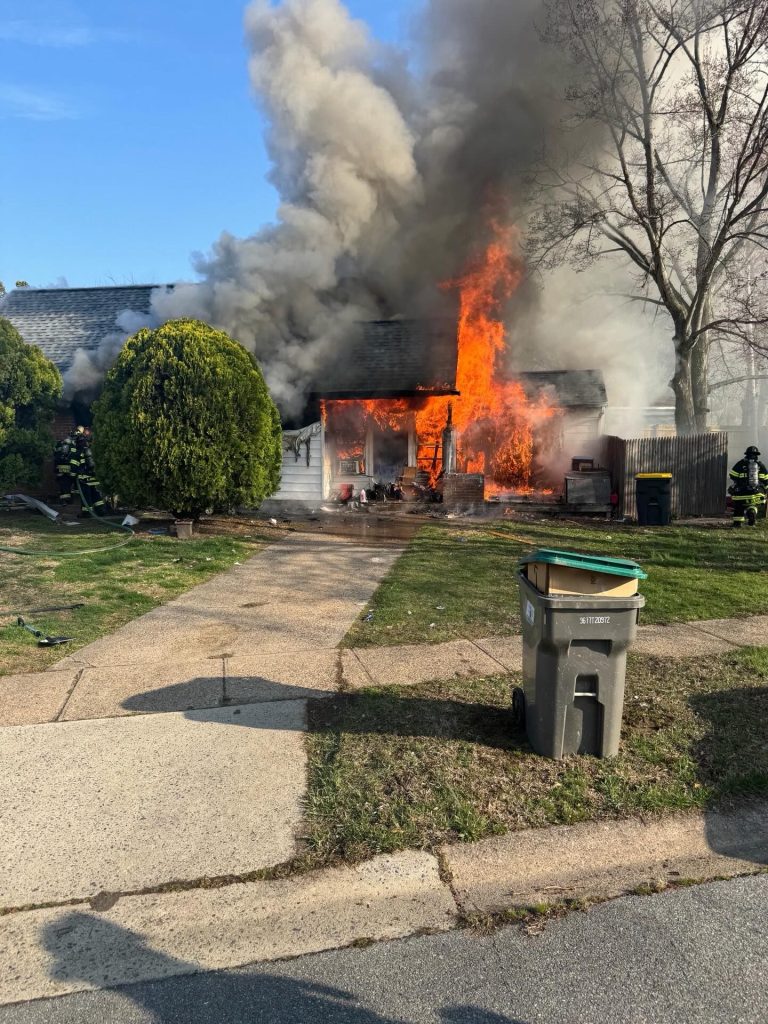 A house is on fire with large flames and thick smoke pouring from the front entrance. Firefighters are present in the yard, and a trash bin is on the sidewalk at the edge of the driveway.